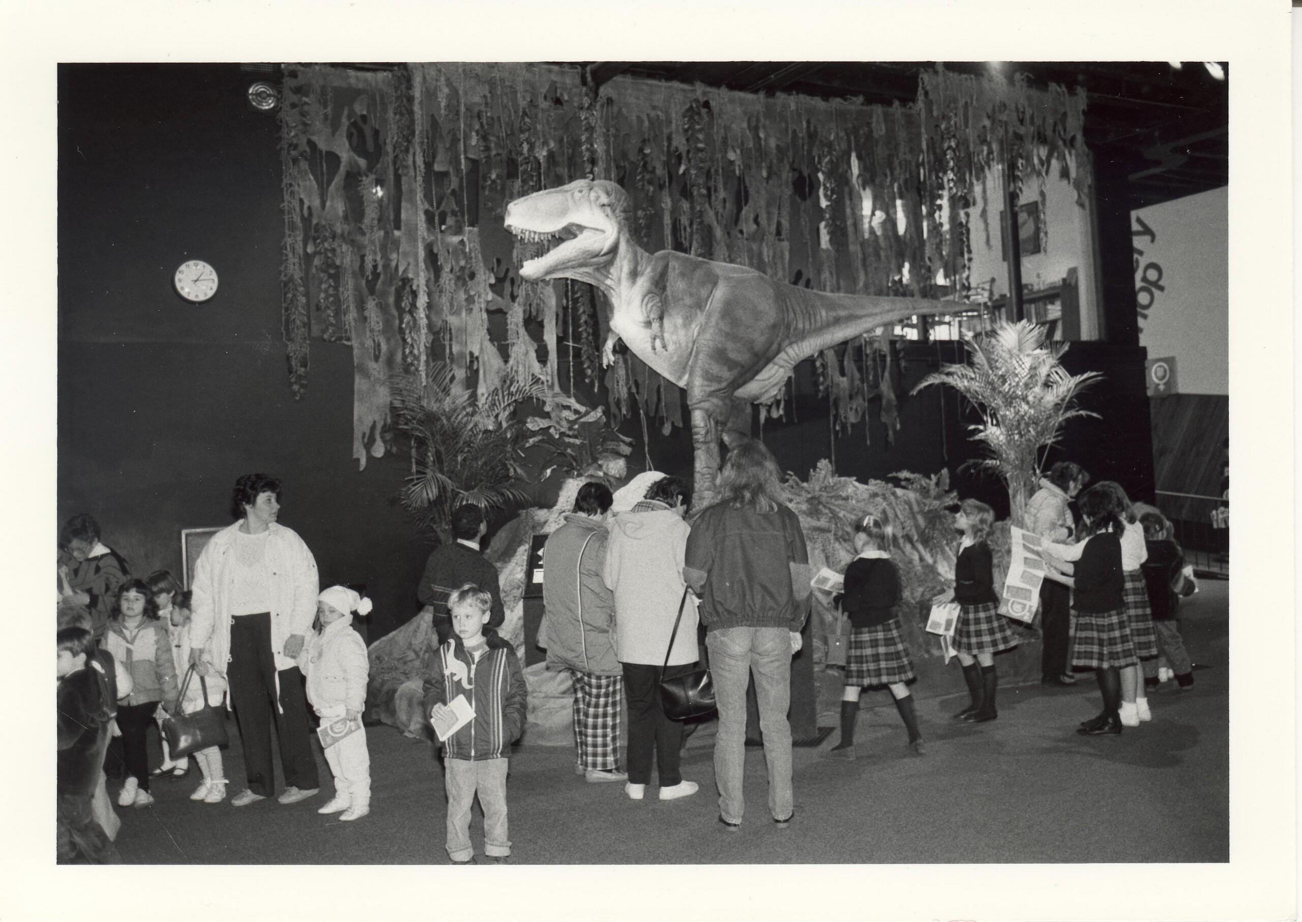 This photo is in black and white. A group of kids with their parents look at dinosaurs at the old COSI building.