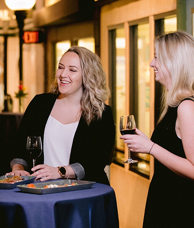 Two smiling women are standing next to a high table holding wine in their hands with appetizers in front of them.